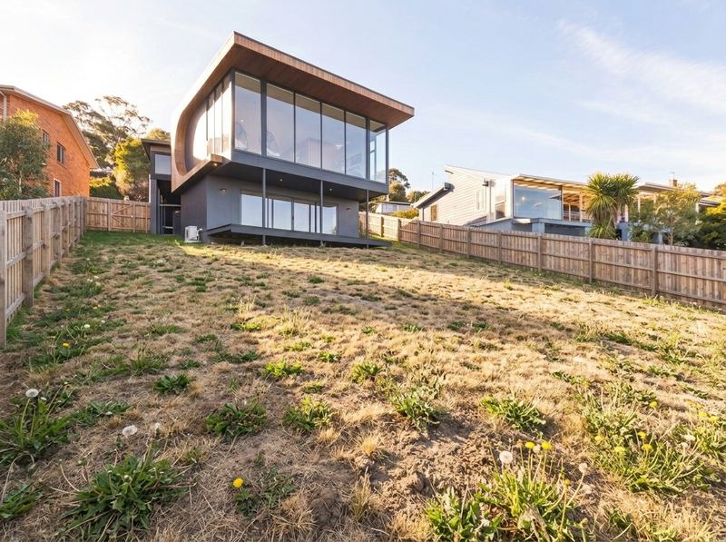 Bare weed-covered yard behind a modern timber and glass home, before landscaping
